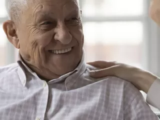 A patient in his home smiles at a healthcare professional, who has her hand placed on the patient's shoulder